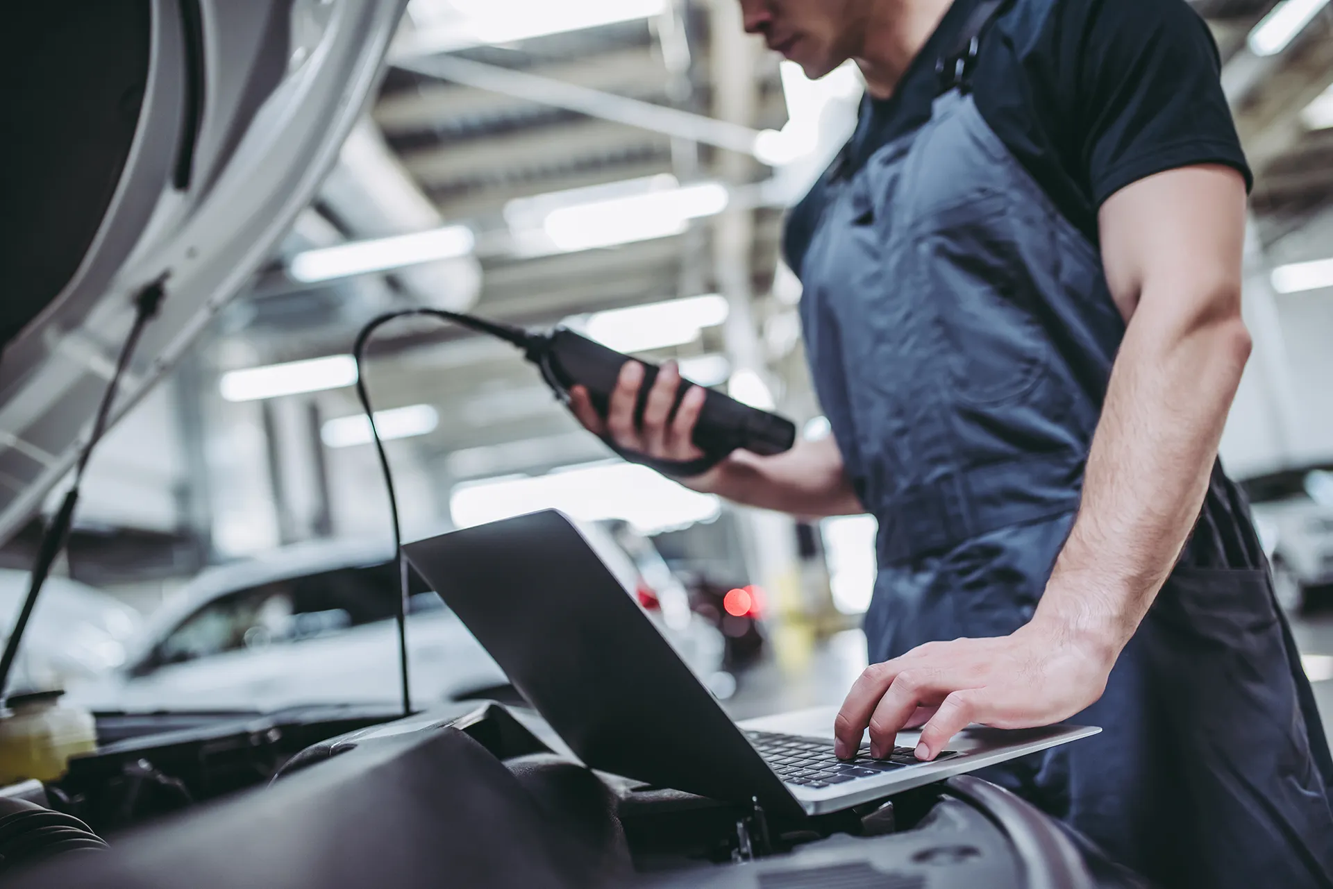 Blurred background shot of a mechanic using a diagnostic tool and a laptop to work on an open car engine.